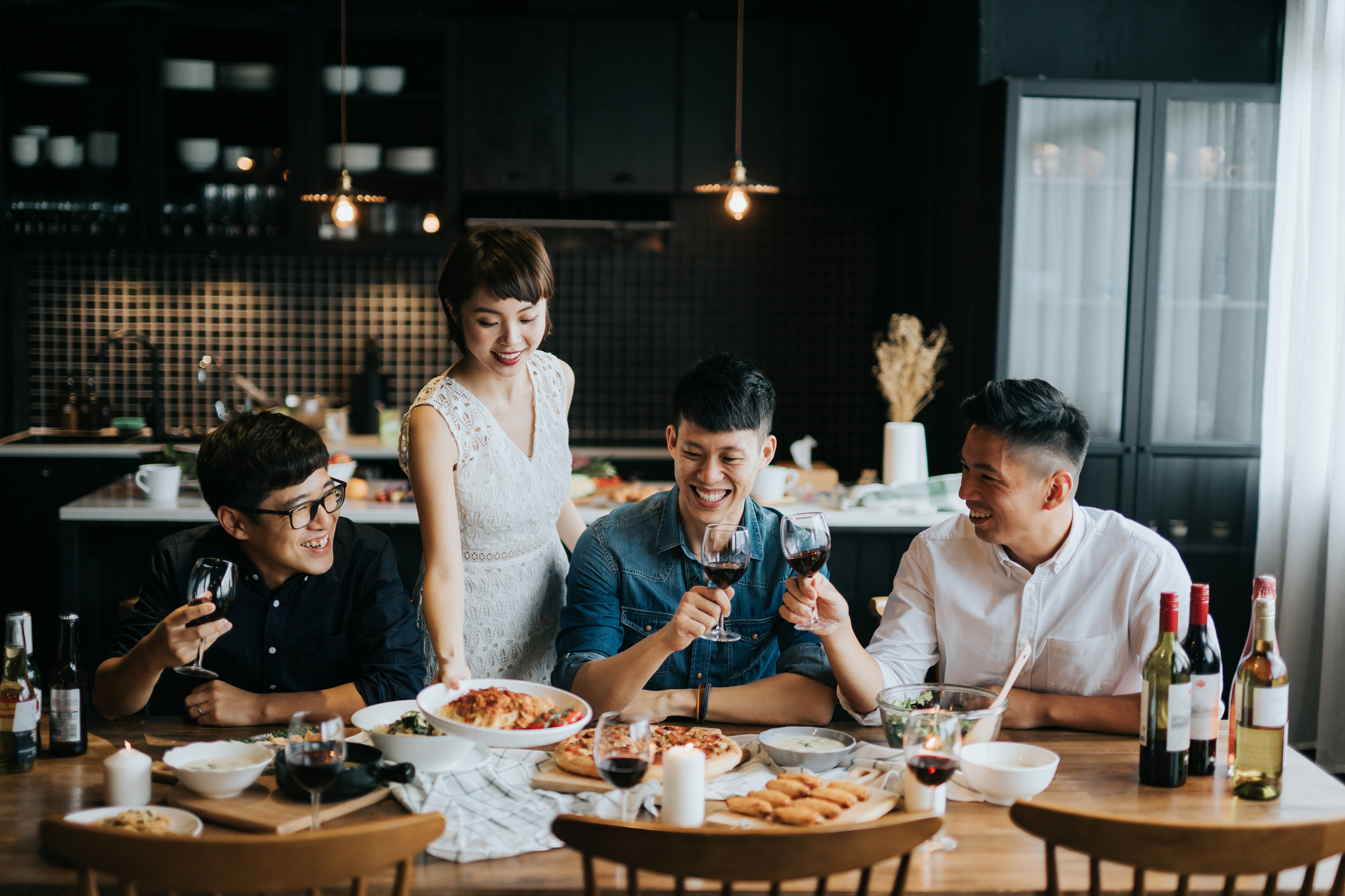 Smiling young Asian woman serving food to friends during party at home, they are smiling joyfully and toasting with red wine