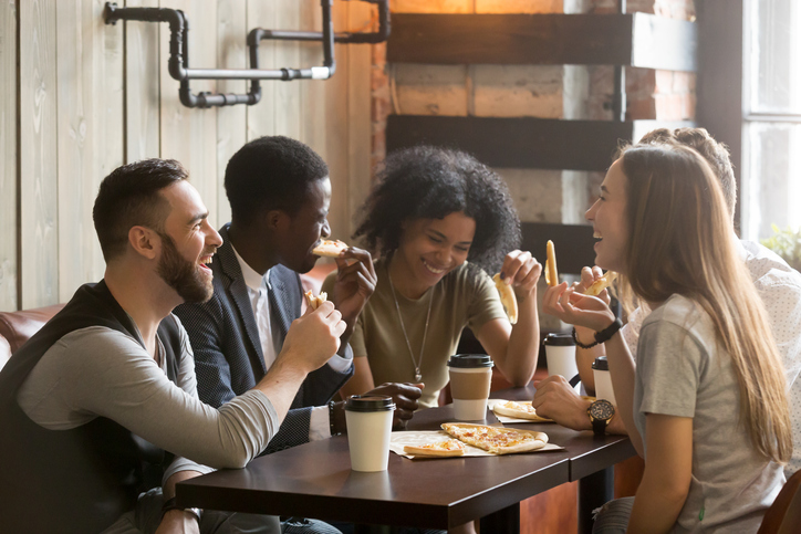 Multiracial happy young people laughing eating pizza together in pizzeria